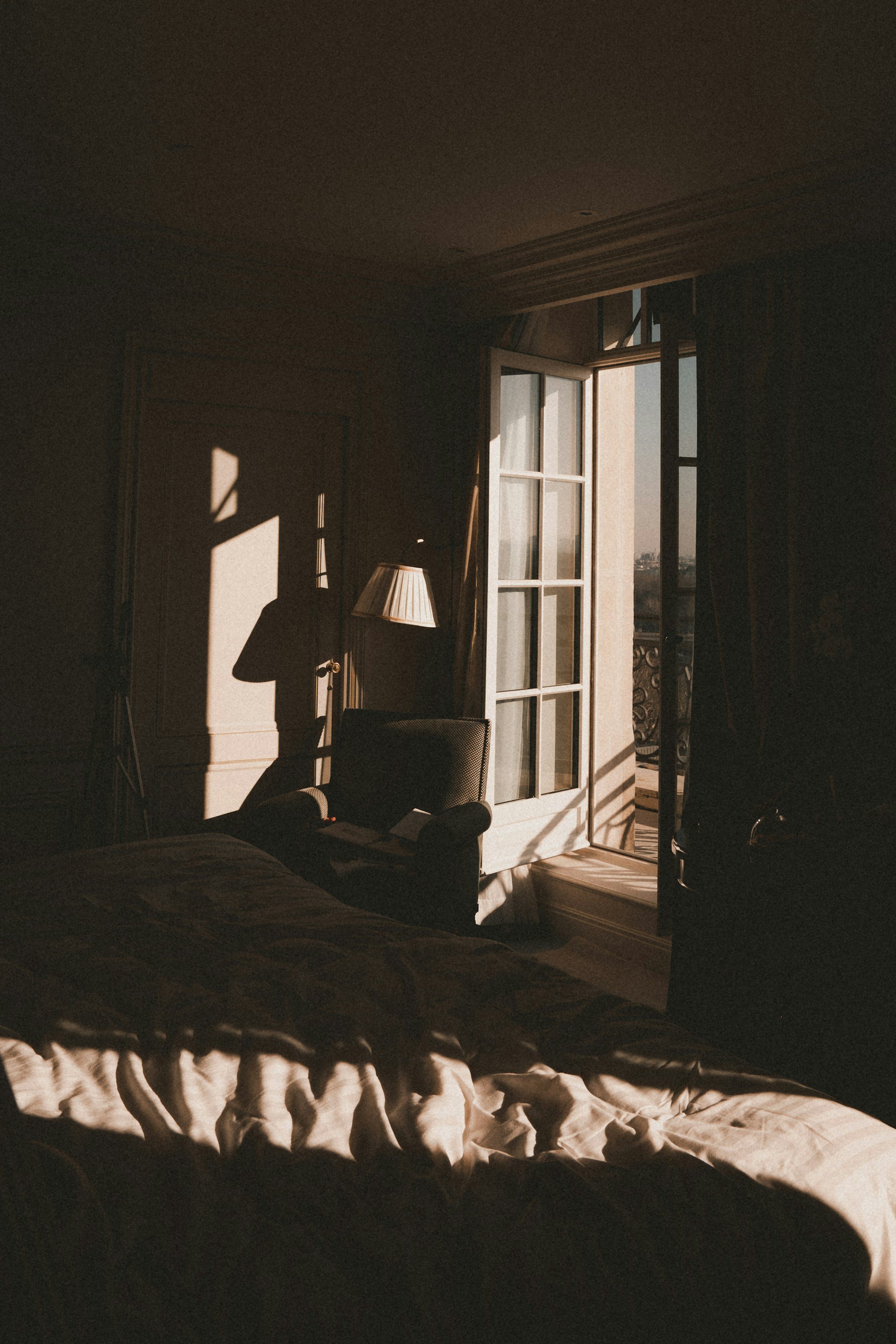 Wedding Website Design, Digital Wedding Stationery, Web Design and Branding for Wedding Professionals | Avelã White. Dimly lit room with an open window, casting shadows on a bed, chair, and floor lamp.