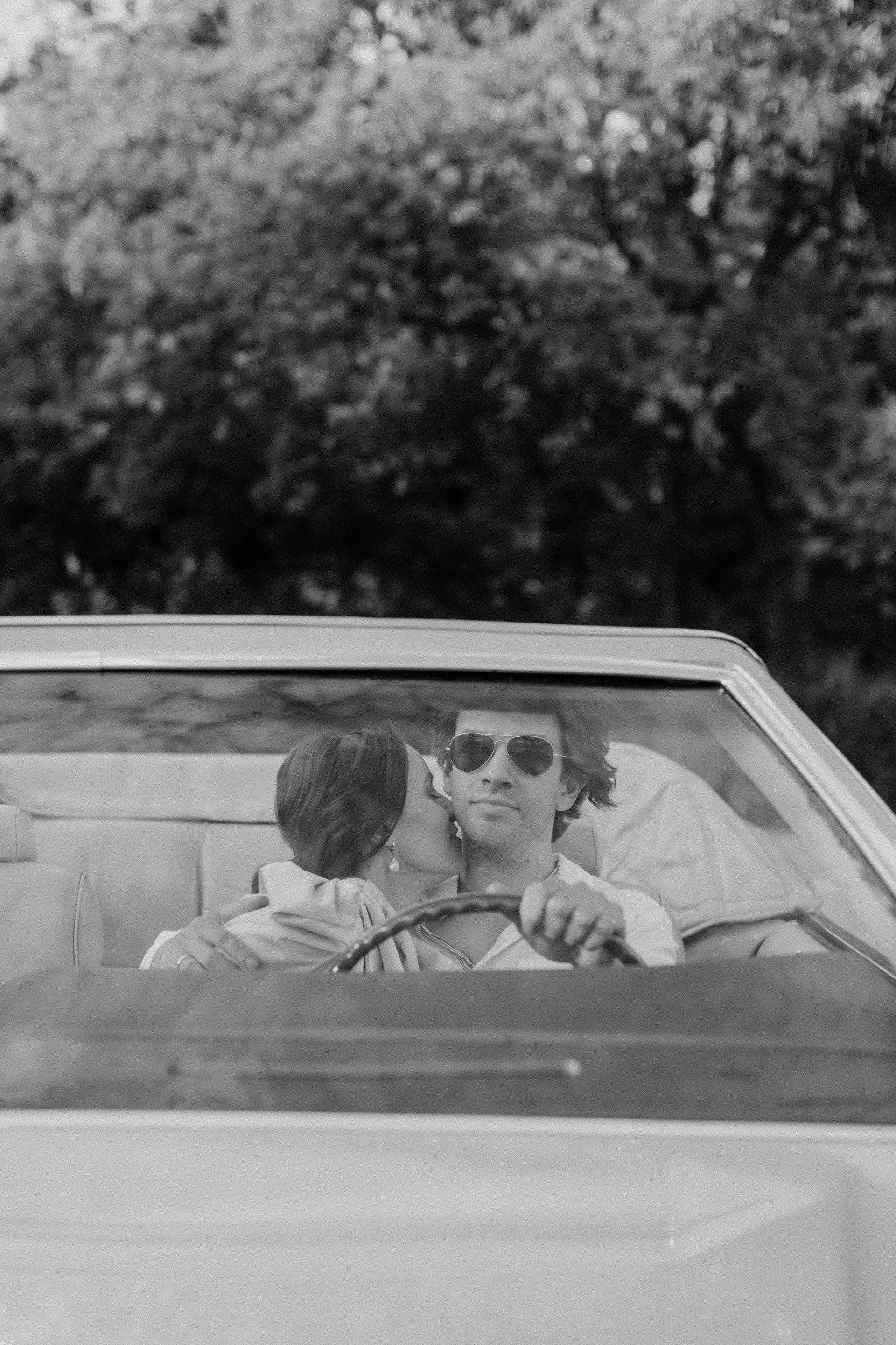 Wedding Website Design, Digital Wedding Stationery, Web Design and Branding for Wedding Professionals | Avelã White. Black and white photo of a couple in a convertible car, with a woman kissing a man who is wearing sunglasses.